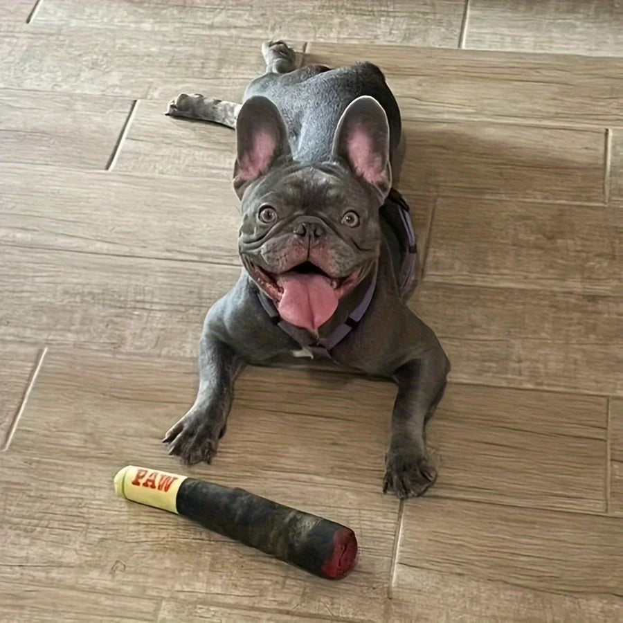 A happy French Bulldog lying on its back on a wooden floor, smiling next to its plush cigar-shaped squeaky toy.