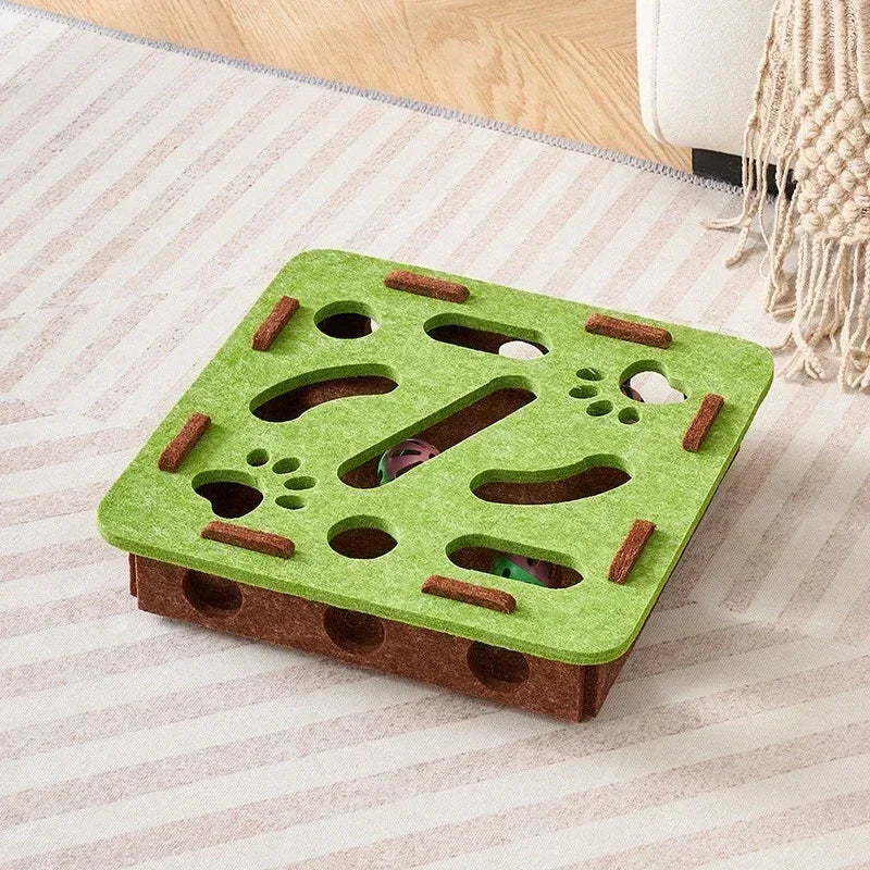 The square-shaped interactive cat puzzle box in a green and brown felt material, placed on a striped rug.