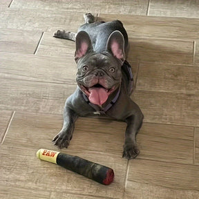 A happy French Bulldog lying on its back on a wooden floor, smiling next to its plush cigar-shaped squeaky toy.