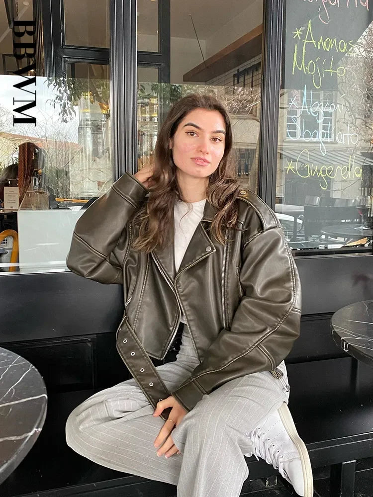 A model sitting outdoors at a cafe wearing the oversized faux leather moto jacket in an olive green color.