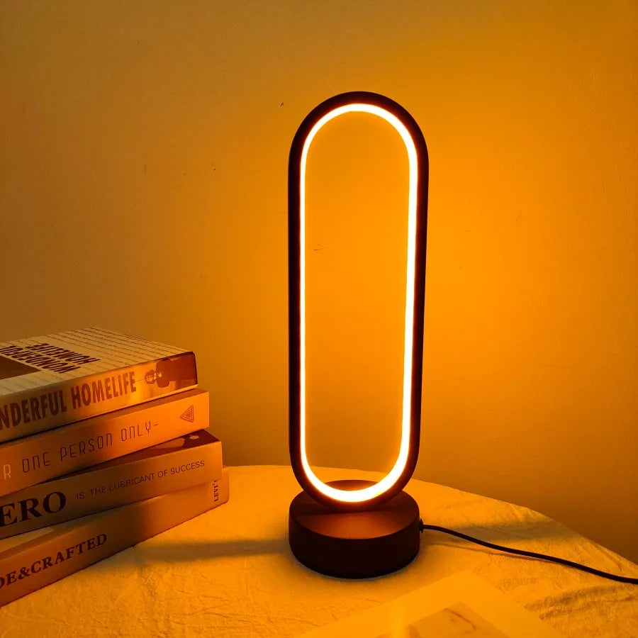 The modern oval LED ring lamp glowing with a warm yellow light on a bedside table next to a stack of books.