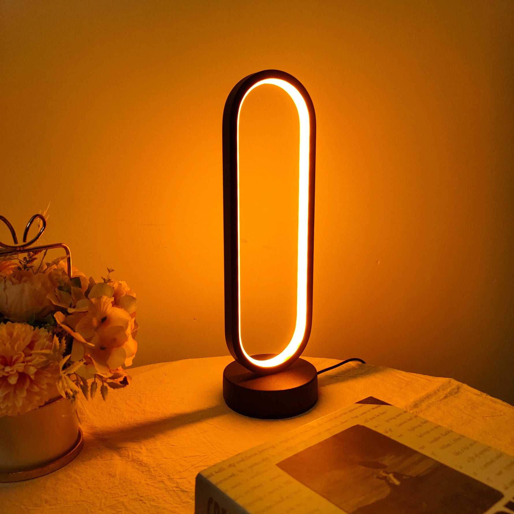 The oval LED desk lamp emitting a warm, inviting glow next to a vase of flowers.