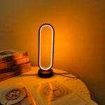 The oval LED table lamp creating a cozy ambient glow on a table with books and a magazine.