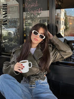 A stylish model with sunglasses holding a coffee cup while wearing the olive green oversized faux leather moto jacket.